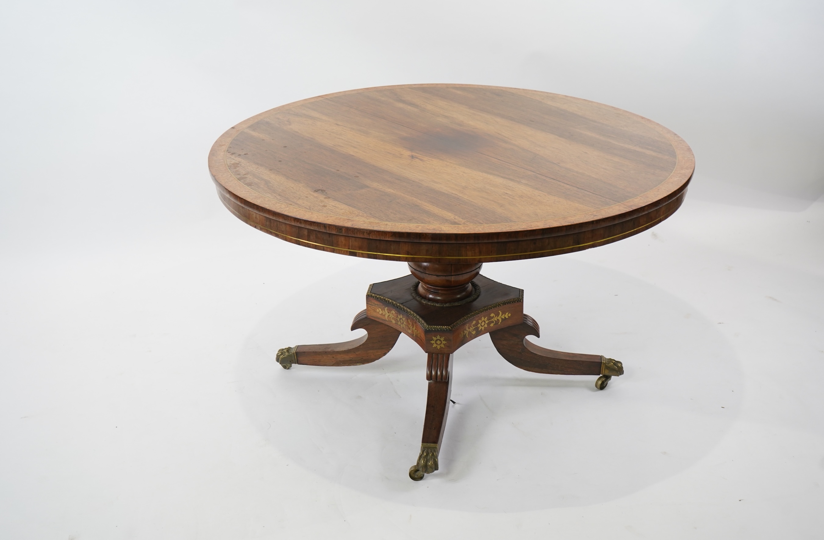 A Regency cut brass inlaid and gilt metal mounted circular rosewood breakfast table, the bird's-eye maple banded tilt top on baluster column, concave platform and quadruple downswept base with cast claw caps and castors,